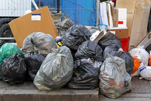 A collection of black and grey plastic rubbish bags filled with discarded waste, piled on a paved area outside a building, with some bags leaning against a metal railing. The bags are tightly knotted and contain various household waste, suggesting a recent rubbish removal process. Surrounding the bags are additional cardboard boxes, some flattened, along with a few loose paper items and other waste materials. The background shows a partially visible blue wall or container, along with a red plastic crate, indicating an outdoor rubbish collection or clearance site possibly in a suburban area near Uxbridge, with local waste disposal regulations in mind. The scene emphasizes the typical appearance of bagged household waste awaiting collection, indicative of a rubbish removal service such as those offered by Uxbridge House Clearance, in a setting consistent with residential or commercial waste disposal in the region.