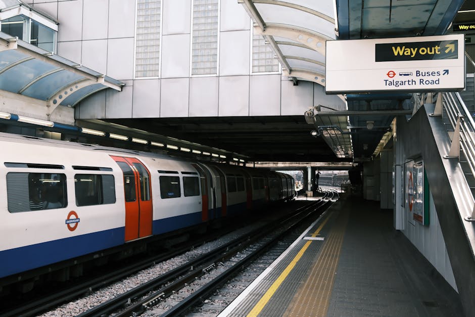 The image shows an underground railway station platform with a train approaching from the distance. The platform surface is covered with beige and brown speckled tiles, and there is a tactile yellow w
